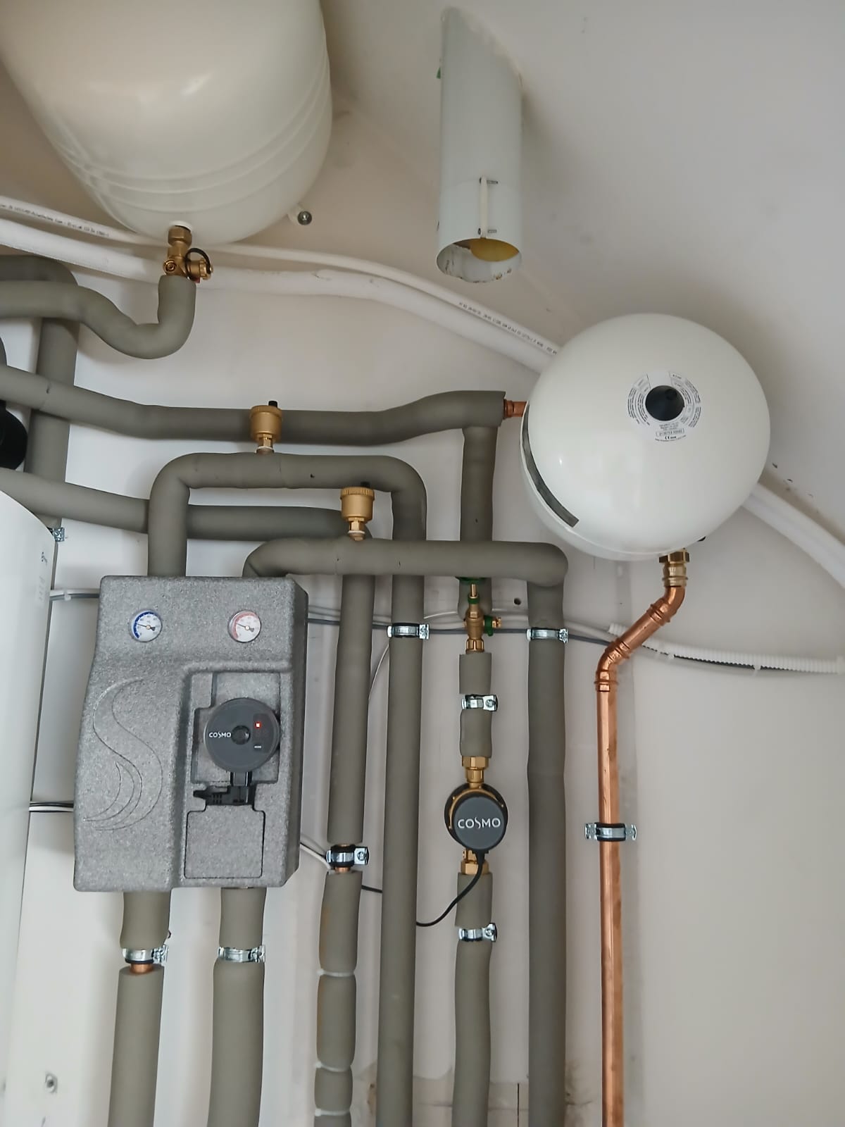 Nahaufnahme von isolierten Heizungsrohren mit zwei weißen Ausdehnungsgefäßen und COSMO-Pumpeneinheit.